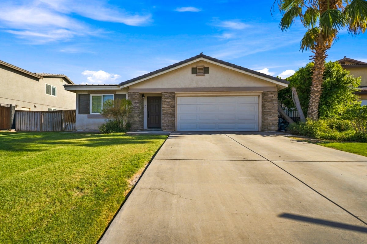 Single-story residential home with white garage door, brick facade, manicured lawn, and driveway on a sunny day