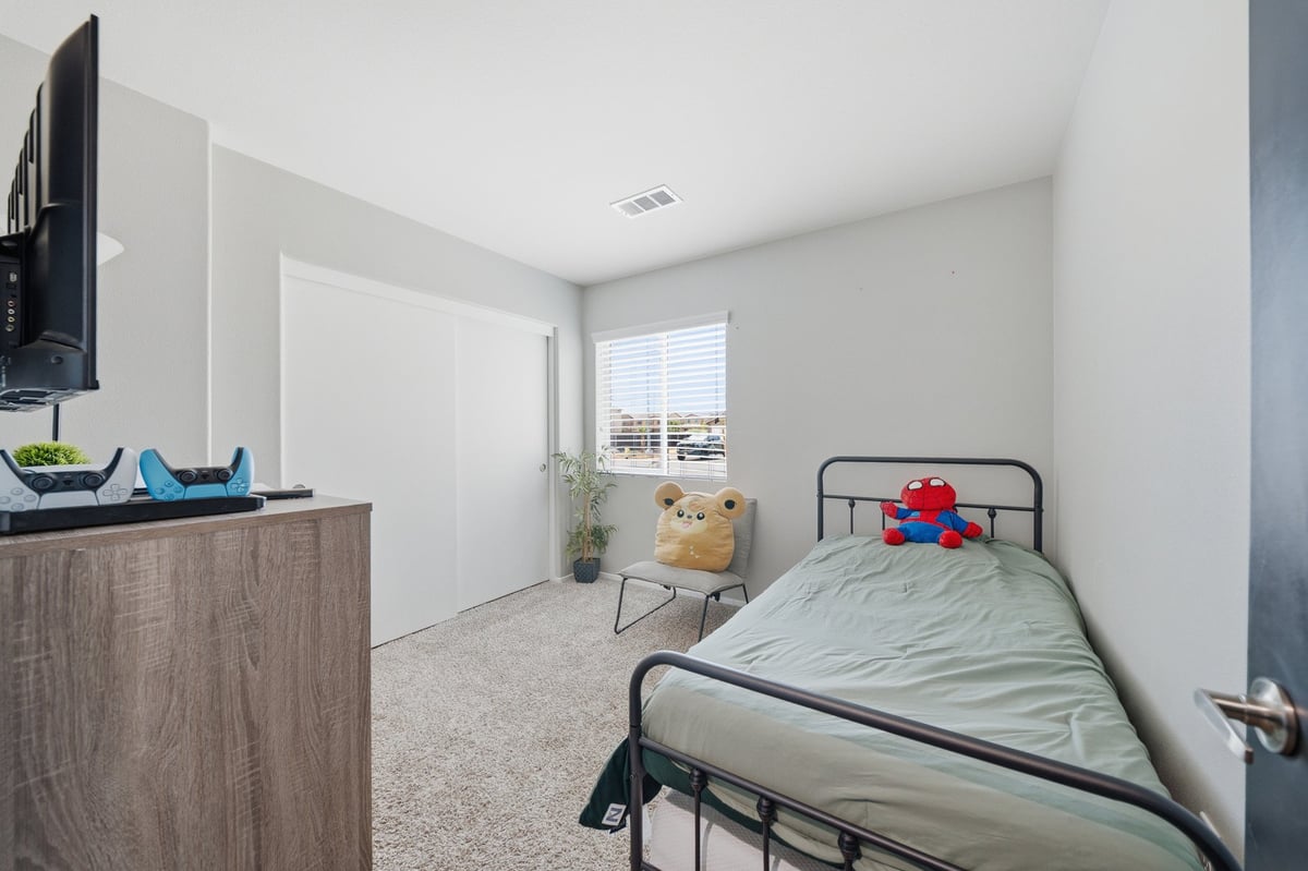 Modern bedroom with metal frame bed, wooden dresser with wall-mounted TV, window with blinds, and stuffed animals on chair