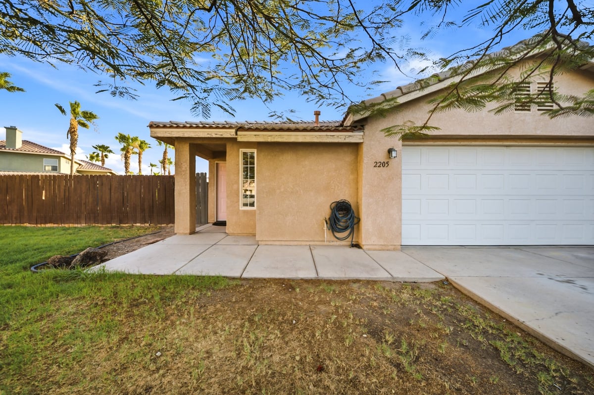 Single-story beige house with white garage, concrete driveway, and landscaped front yard with palm trees