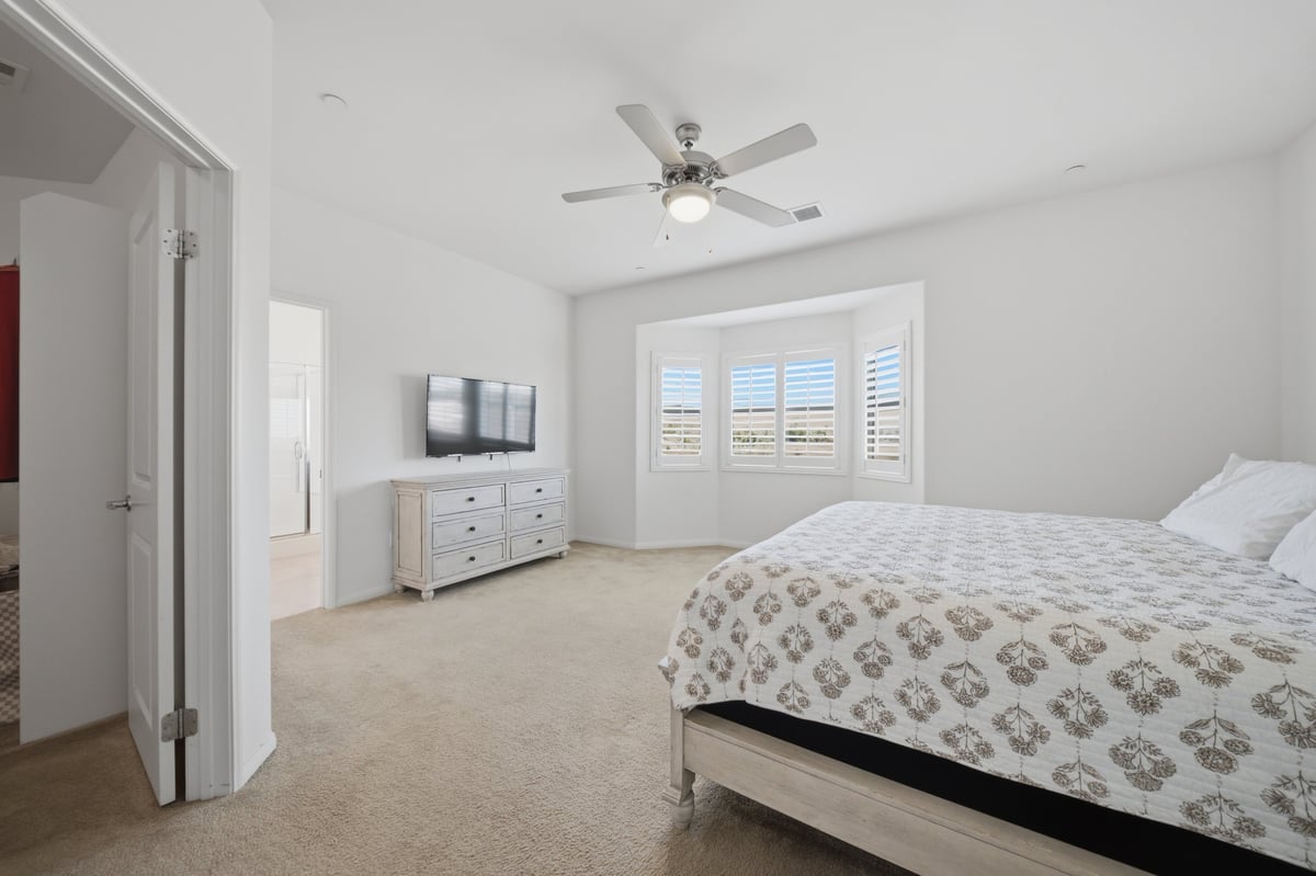 Bright modern bedroom with king bed, white dresser with TV, ceiling fan, and white shuttered doors leading to balcony