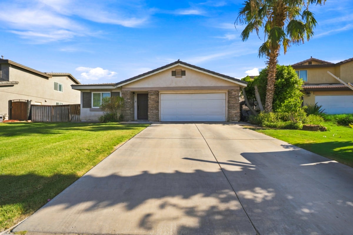 Single-story beige house with white garage, green lawn, palm tree, and clear blue sky