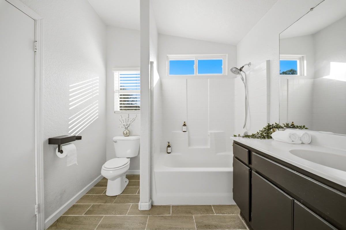 Modern bathroom with white walls, dark vanity, toilet, bathtub with shower enclosure, and tile flooring