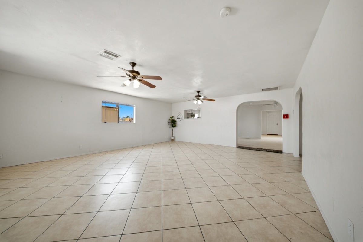 Empty room with beige tile flooring, ceiling fans, bright windows, and white walls in a spacious residential space
