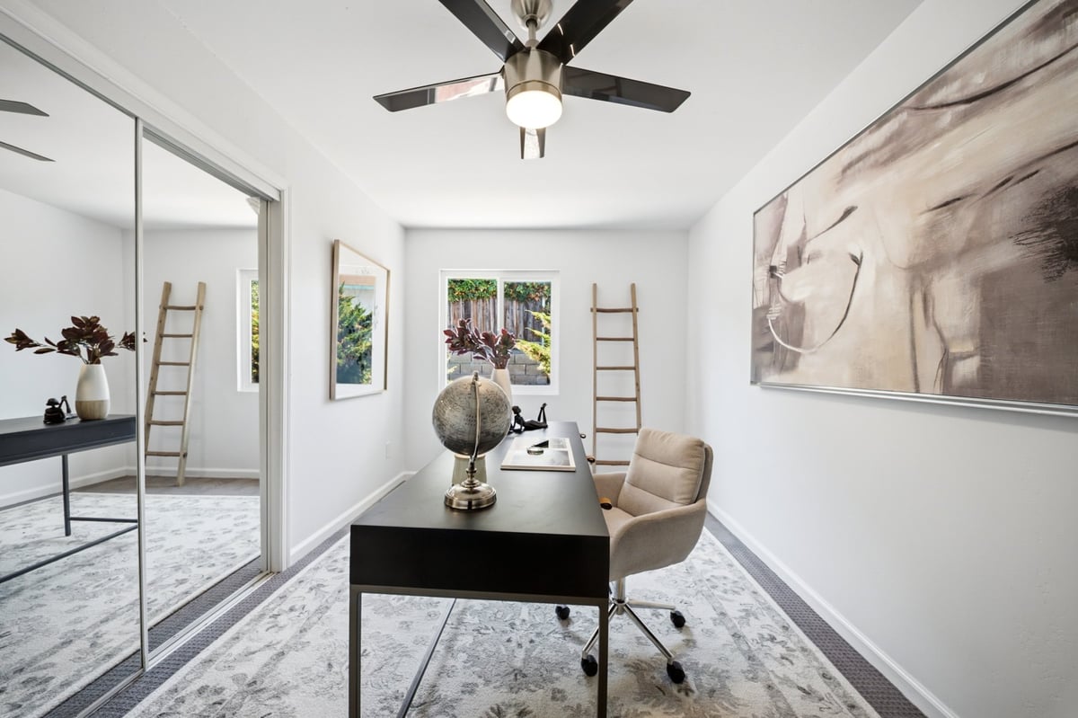 Modern home office with black desk, beige chair, ceiling fan, large mirror, and artwork on white walls