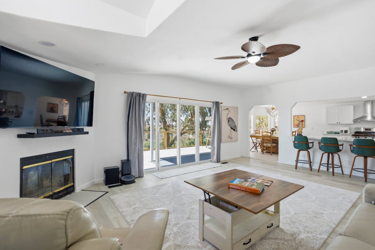 Modern living room with fireplace, ceiling fan, neutral furnishings, glass patio doors, and kitchen visible in background