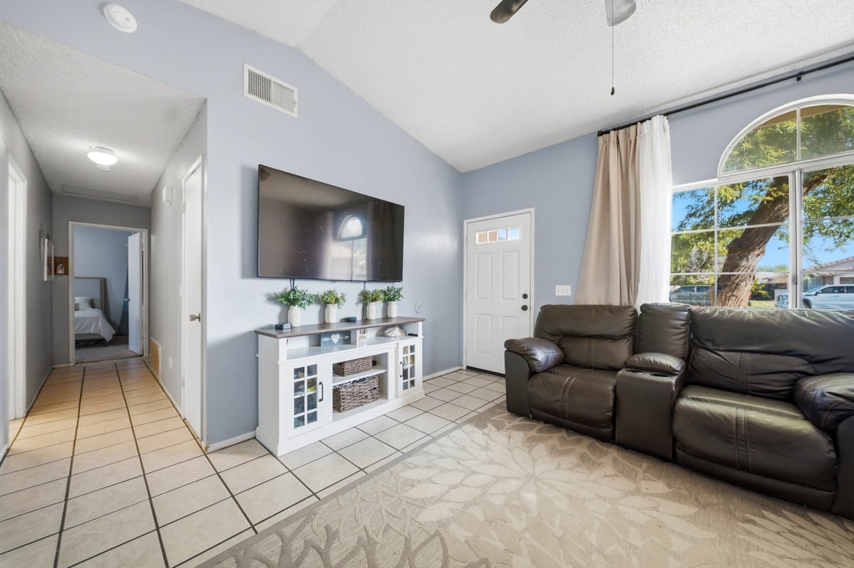 Modern living room with light blue walls, brown leather sectional, wall-mounted TV, white entertainment stand, and large windows overlooking landscaping