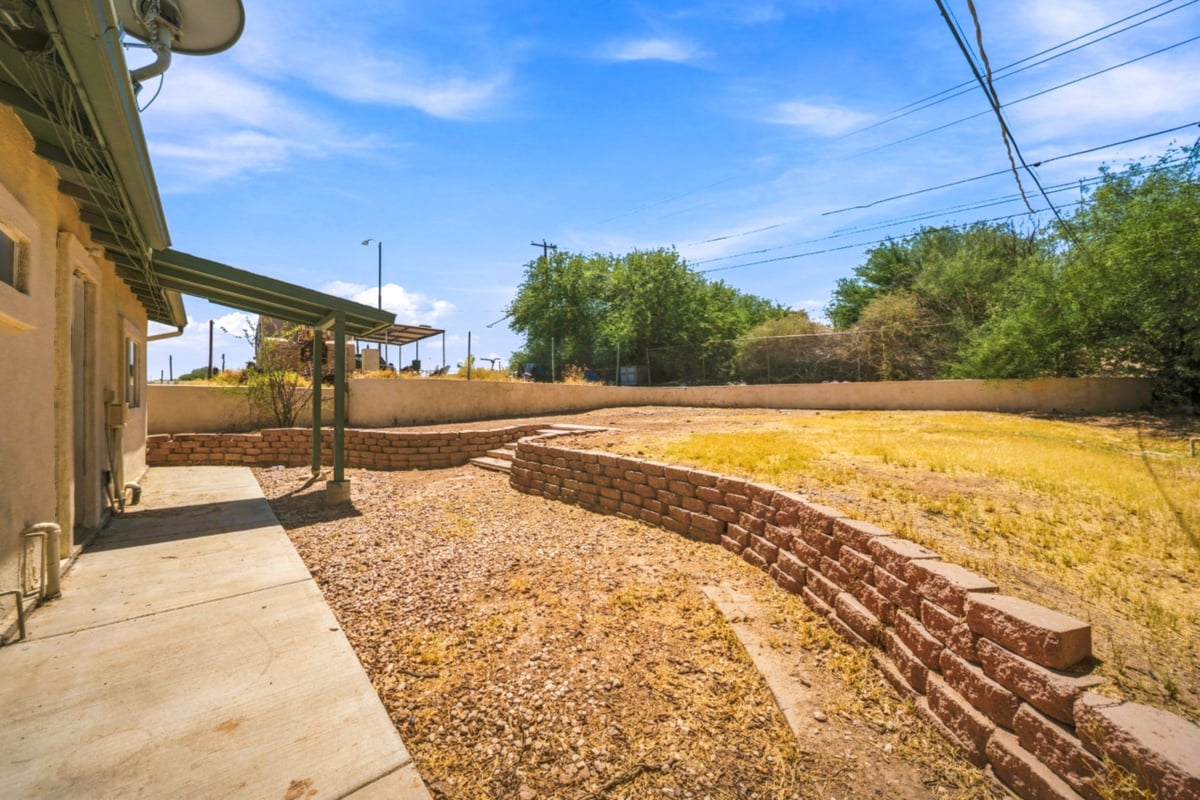Residential backyard with concrete patio, gravel landscape, brick retaining wall, and open dry yard under blue sky