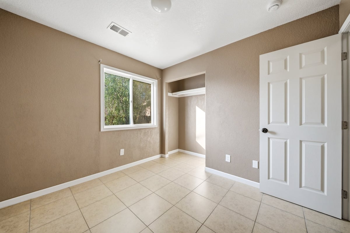 Empty bedroom with tan walls, tile flooring, large window, white closet door, and ceiling light fixtures