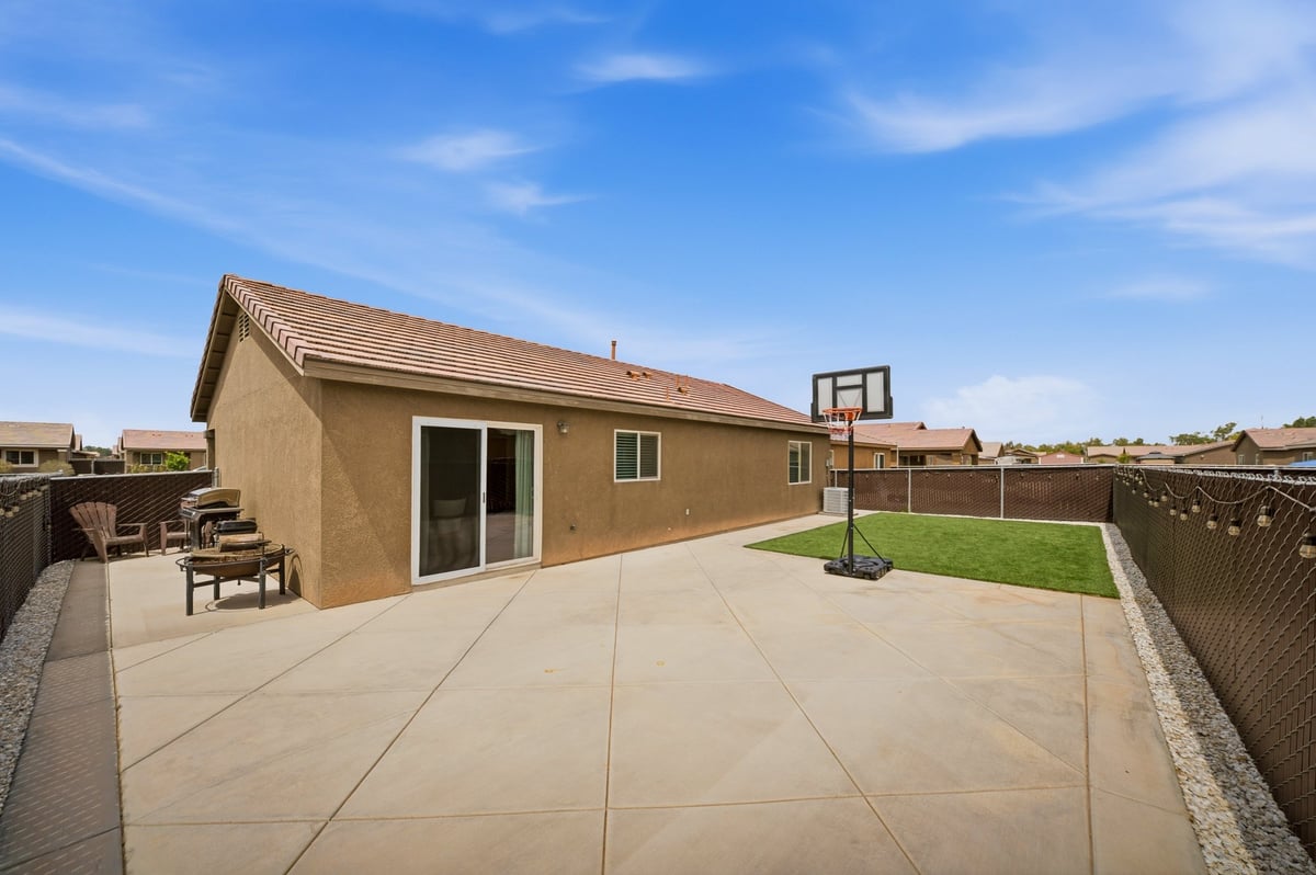Modern tan stucco house with concrete patio, basketball hoop, and fenced backyard under clear blue sky