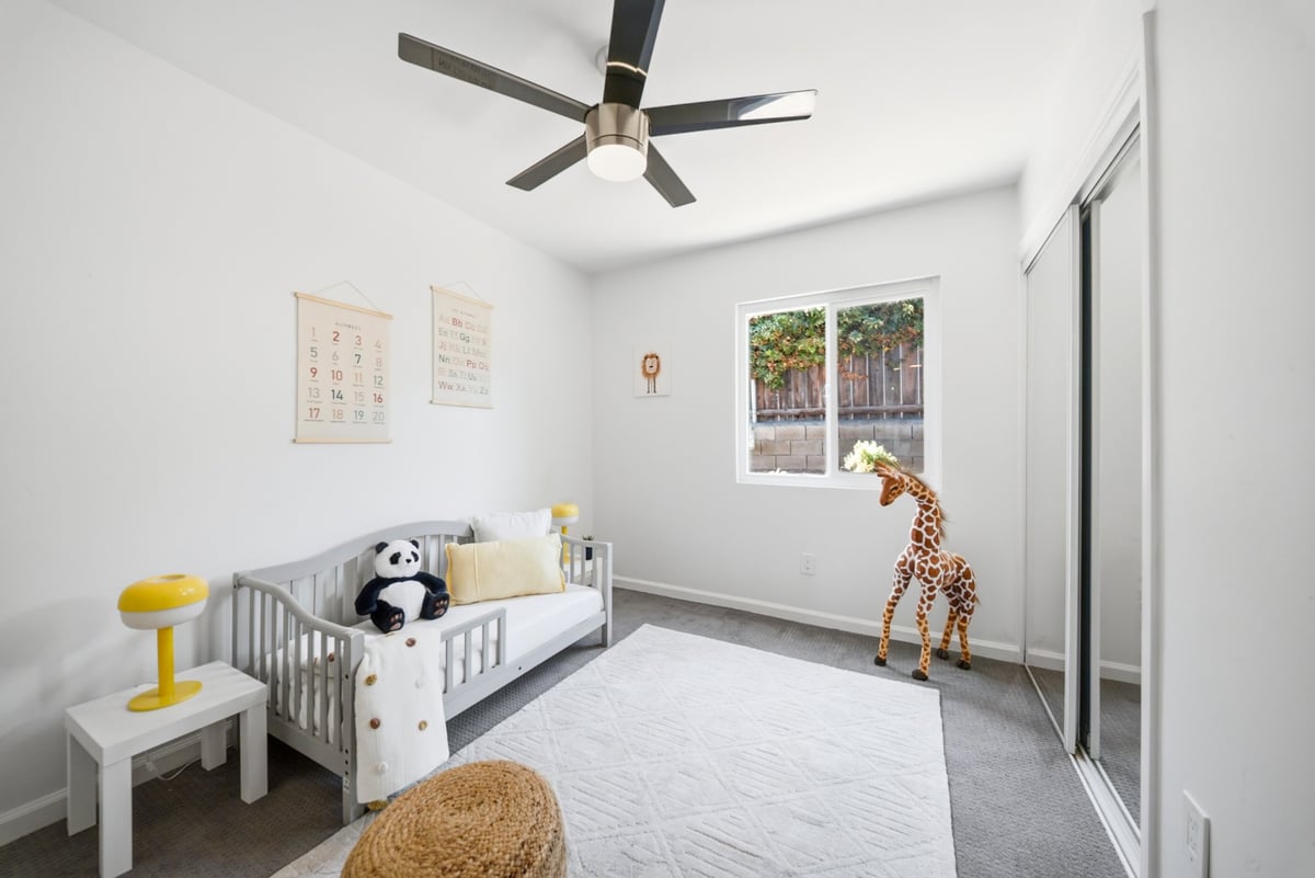 Modern nursery room with gray crib, white nightstand with yellow lamp, ceiling fan, framed artwork, large windows, and stuffed animals on gray flooring