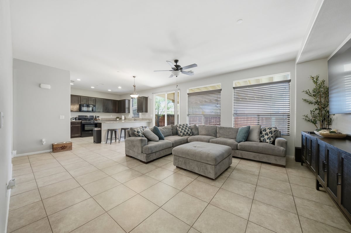 Modern open-concept living room with gray sectional sofa, ottoman, tile flooring, and kitchen area visible in background