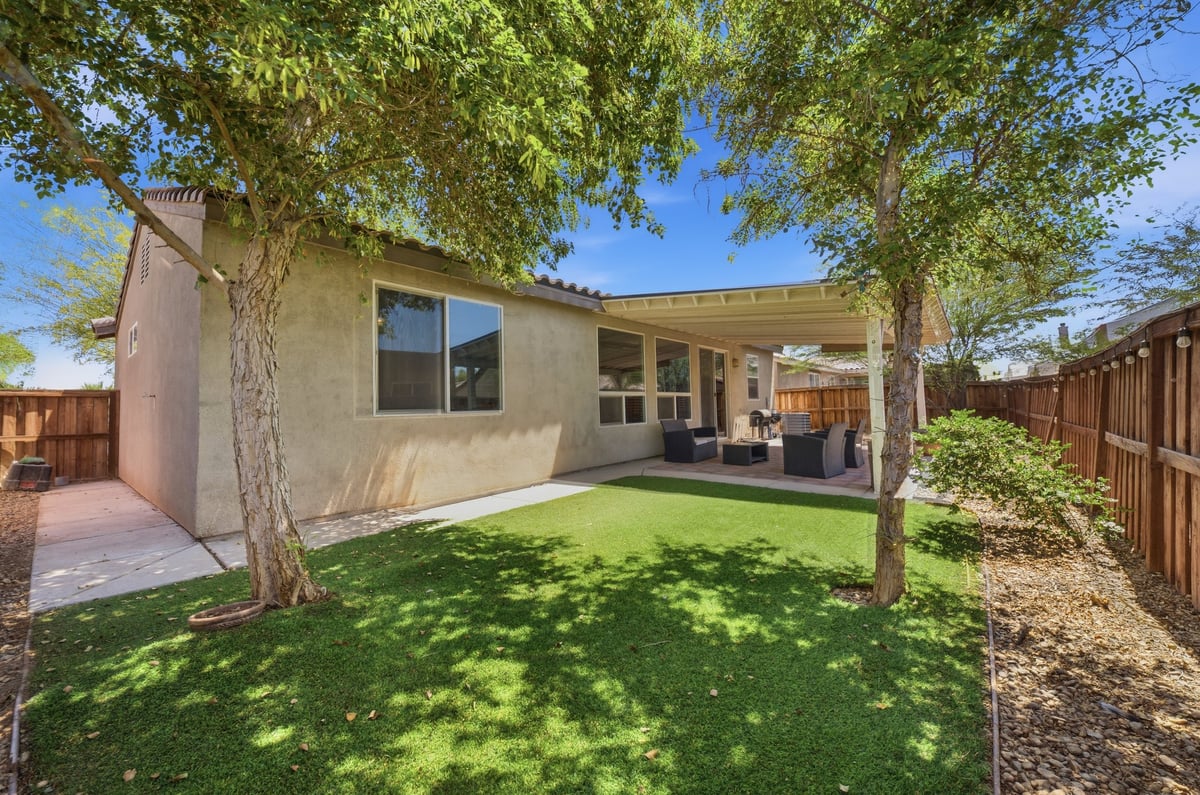 Single-story house with patio and green lawn shaded by mature trees, wooden fence visible on right side