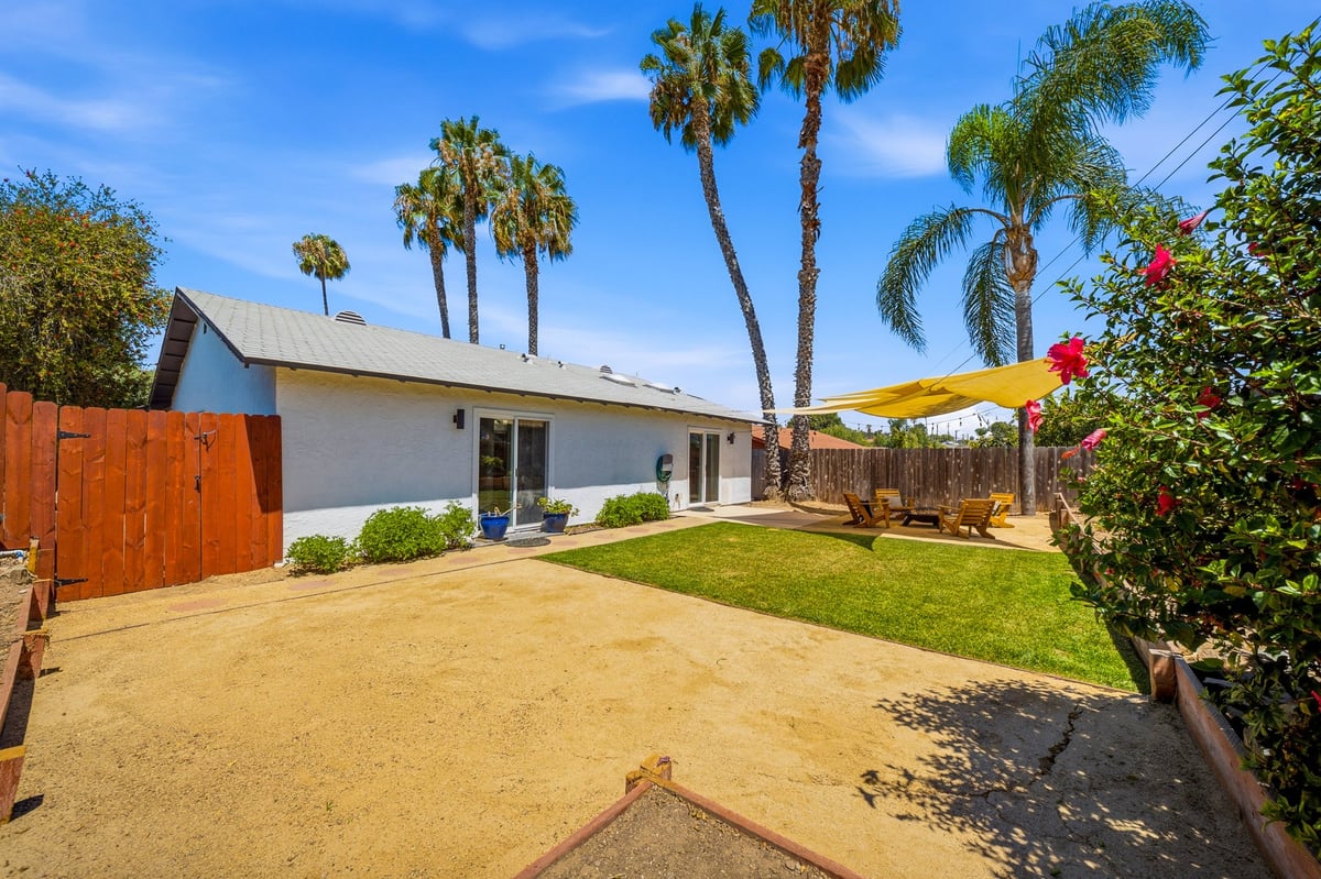 Single-story white house with palm trees, sandy yard, green lawn, yellow patio umbrella, and wooden fence on a sunny day