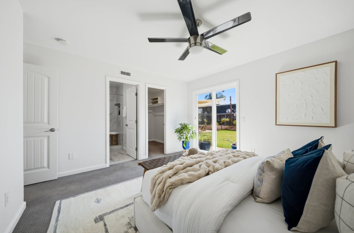 Modern bright bedroom with white bedding, dark ceiling fan, glass doors opening to patio, and neutral decor