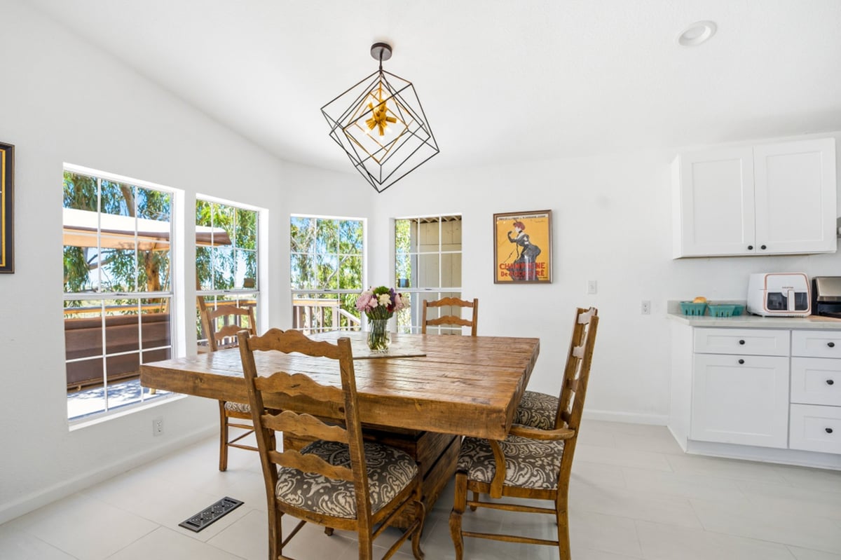 Bright dining room with wooden table, upholstered chairs, white cabinets, large windows, and geometric ceiling pendant light