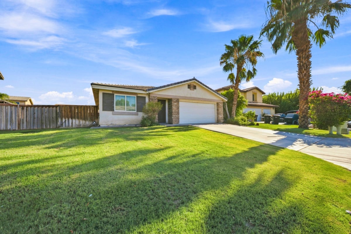 Single-story residential home with beige exterior, white garage, and manicured green lawn under clear blue sky with palm trees