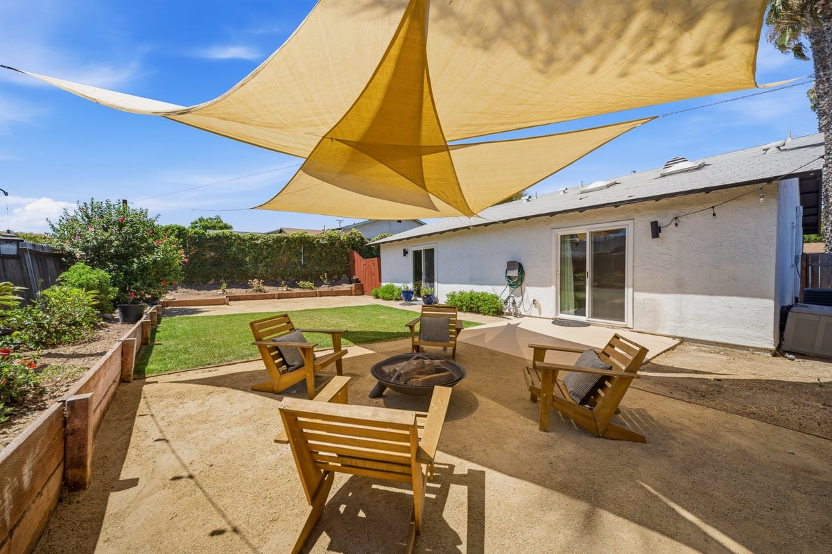 Backyard patio with golden shade sail, wooden chairs around fire pit, white house, and green lawn under blue sky