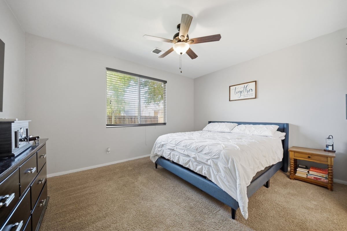 Bright bedroom with queen-size bed, ceiling fan, window with blinds, dresser, and beige carpet throughout.