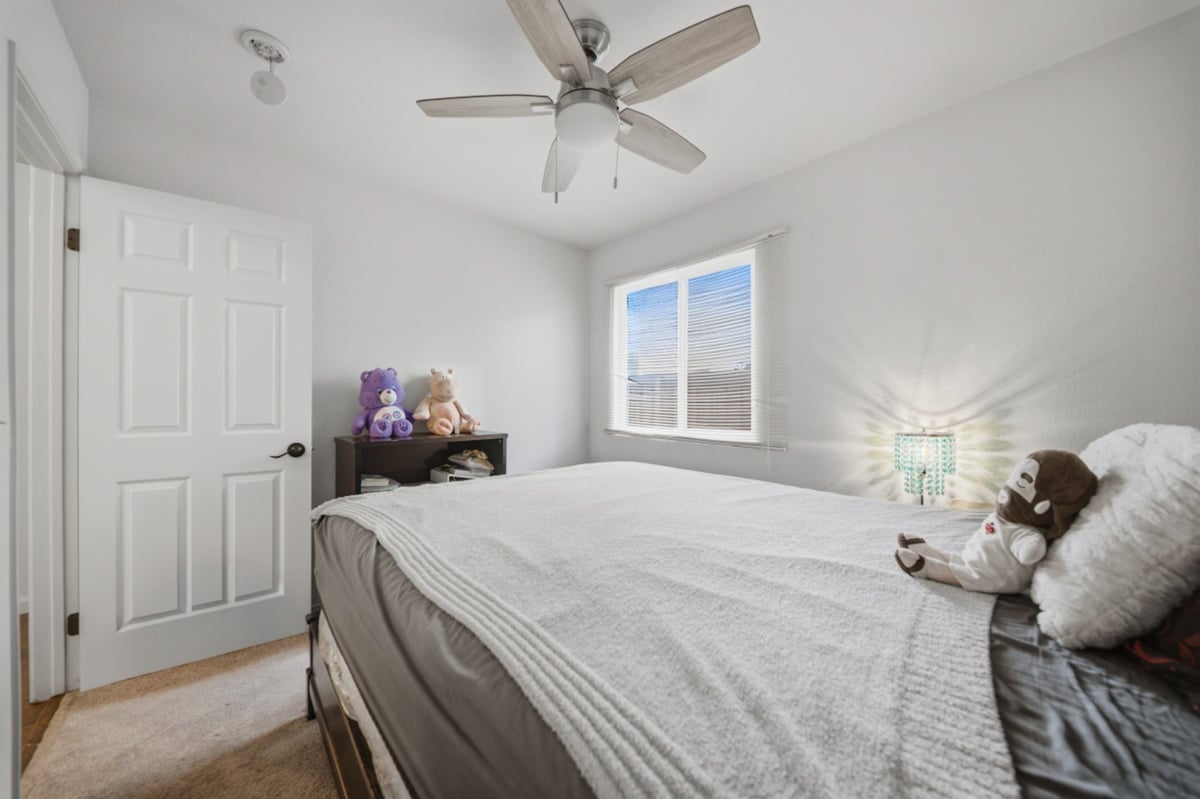 Bright modern bedroom with queen bed, ceiling fan, large window with mountain view, white closet doors, and purple decorative items on floating shelf
