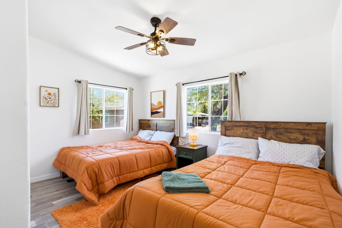 Bright bedroom with two beds with orange comforters, wooden headboards, ceiling fan, and large windows overlooking greenery