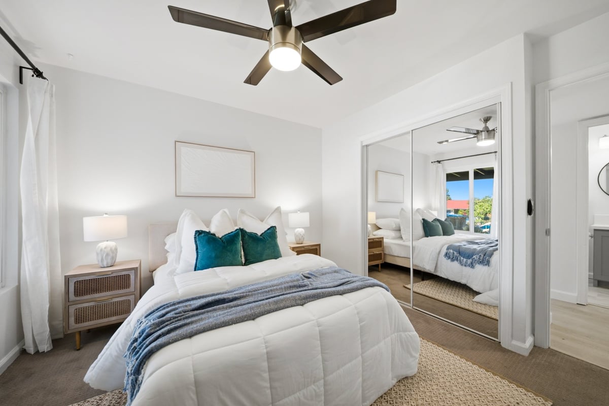 Modern bedroom with white bedding, teal pillows, ceiling fan, mirrored closet doors, and coastal window views