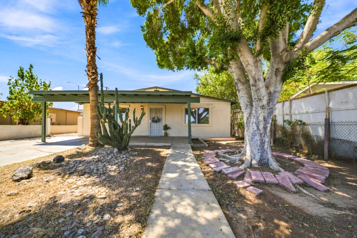 Single-story beige house with green roof shaded by large mature tree, concrete walkway leading to entrance, fenced yard