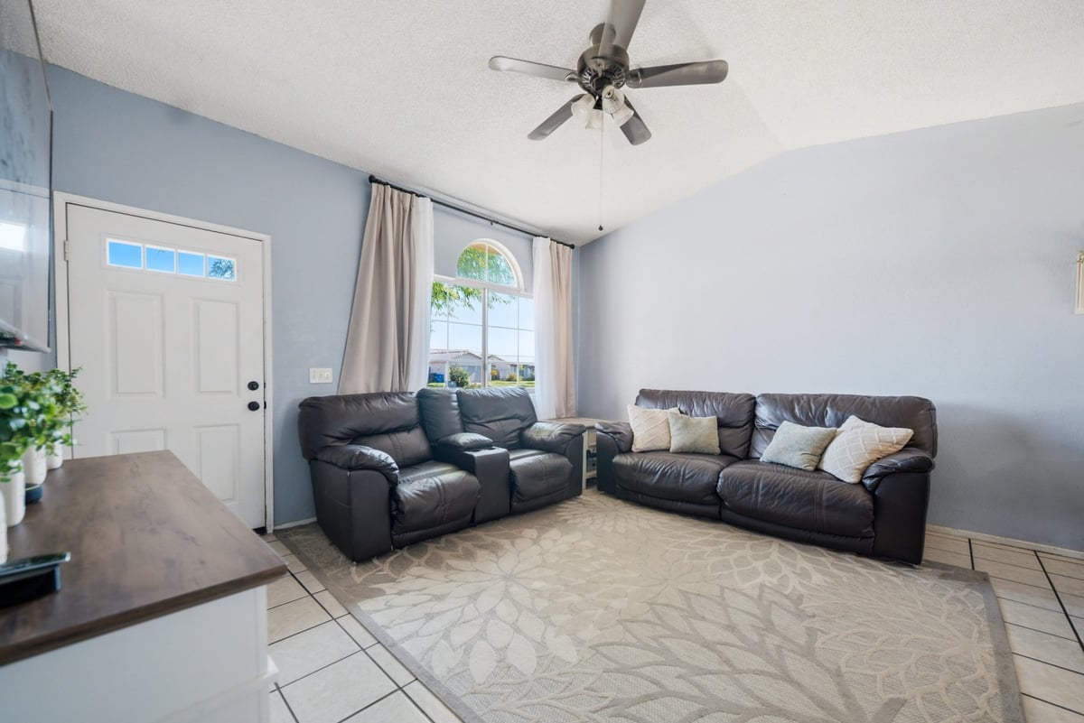 Spacious living room with black leather sectional sofa, ceiling fan, large windows with curtains, white front door, and light tile flooring