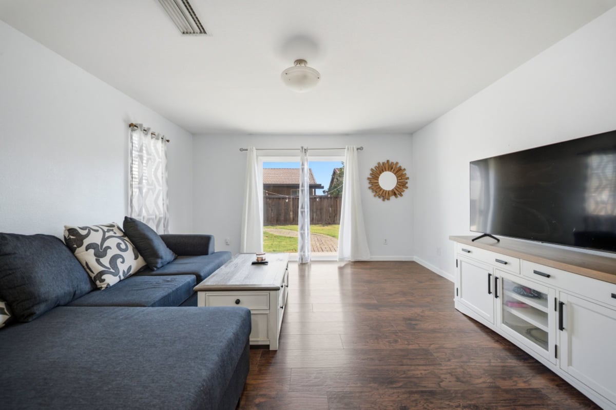 Bright modern living room with blue sectional sofa, white media console, wooden floors, and glass doors leading to outdoor patio