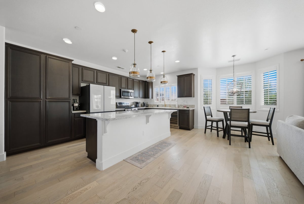 Modern open-concept kitchen with dark cabinets, white island, stainless steel appliances, and dining area with light wood flooring