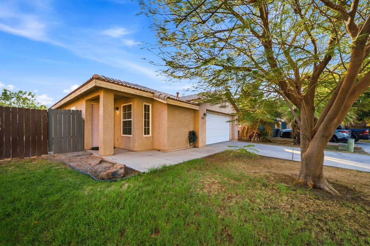 Single-story tan stucco house with white garage, green lawn, large tree, and clear blue sky