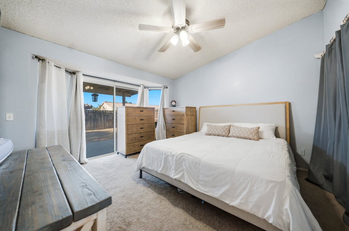 Spacious modern bedroom with light blue walls, queen bed with white linens, wooden nightstands, ceiling fan, and glass patio doors leading to backyard
