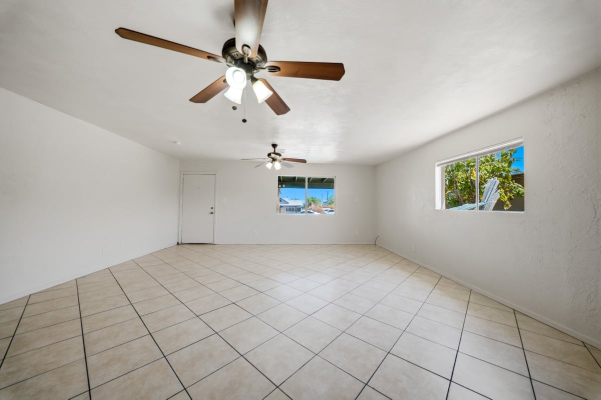 Empty room with beige tile flooring, white walls, ceiling fan with brown blades, and two windows with natural light