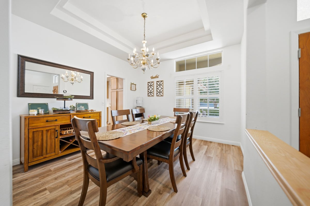 Bright dining room with wooden table, chairs, and buffet cabinet, featuring a crystal chandelier and large windows with shutters