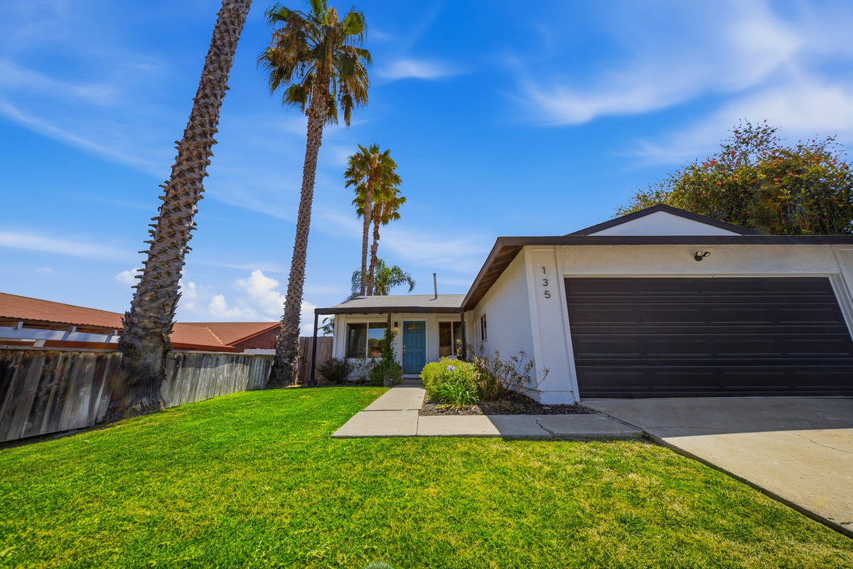 Single-story house with white exterior, dark garage door, and manicured front lawn with palm trees under blue sky