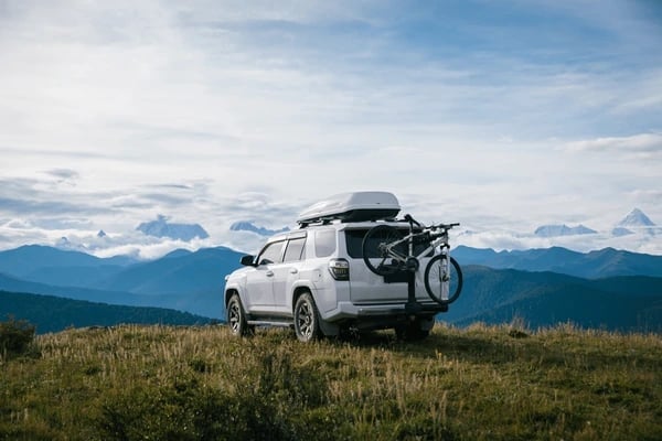 White SUV with roof rack and mountain bikes parked on grassy hillside overlooking snow-capped mountains