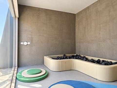 Modern childrens sensory play room with curved cream-colored seating filled with black balls, green and blue circular mats, and large windows