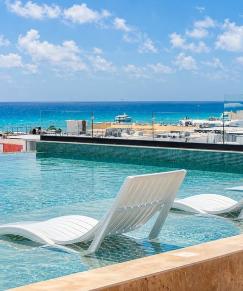 Modern white lounge chairs in a turquoise pool overlooking a marina with boats and clear blue ocean