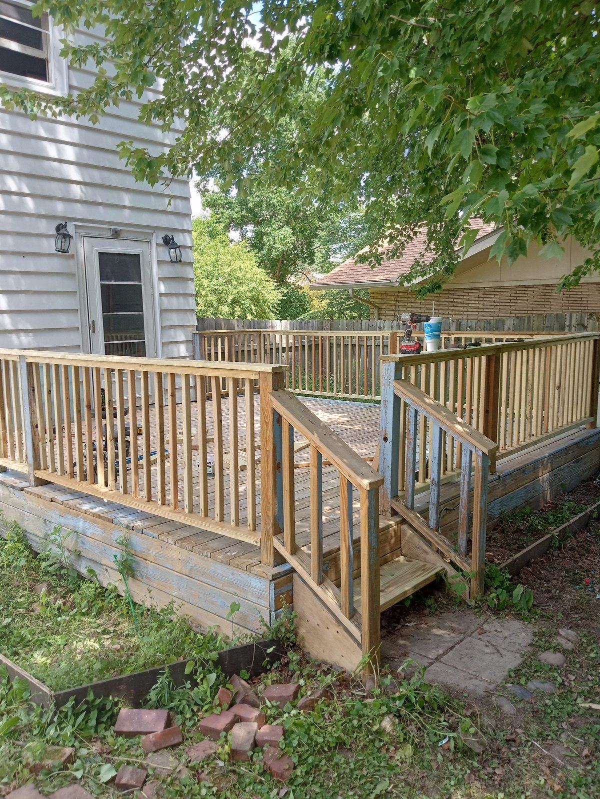 Wooden deck with railings connecting two residential houses, surrounded by green trees and landscaping