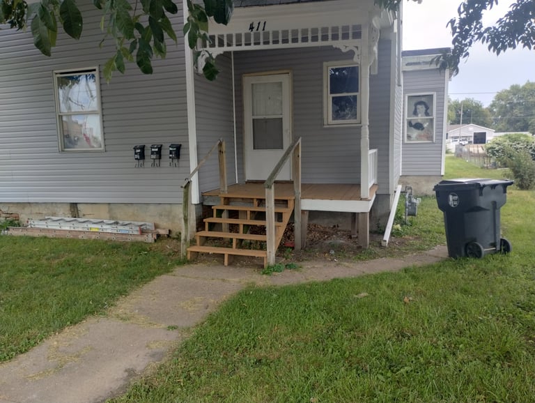 Front entrance of a mobile home with wooden steps, gray door, and black trash bin on grassy lot