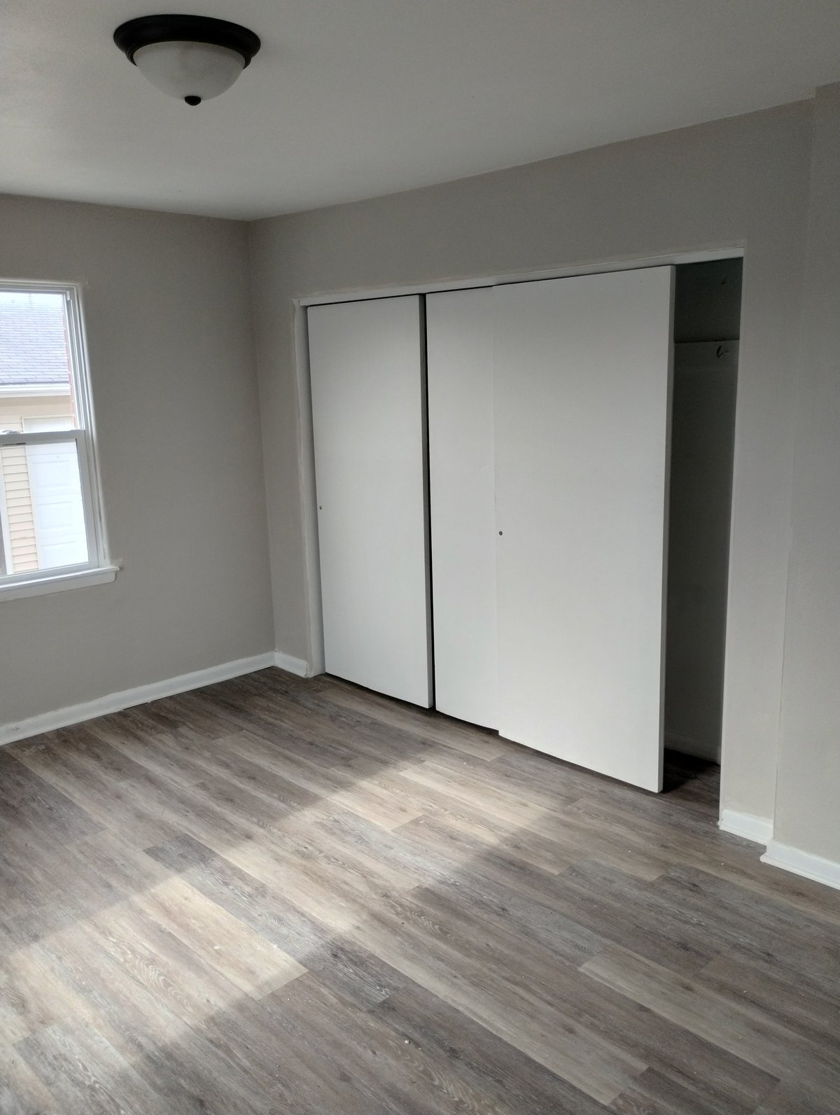 Empty bedroom with white sliding closet doors, gray walls, wood-grain flooring, window, and ceiling light fixture