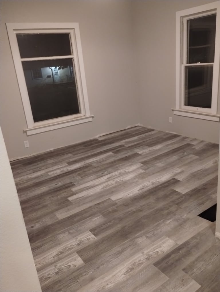 Empty room with gray wood-look laminate flooring, white walls, two windows with dark frames, and white electrical outlets