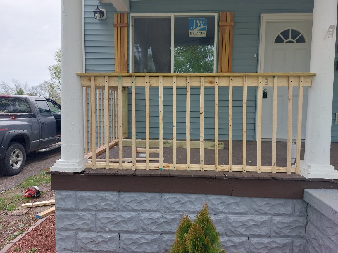 Finished wooden porch railing on elevated porch of light blue house with gray stone foundation and landscaping below