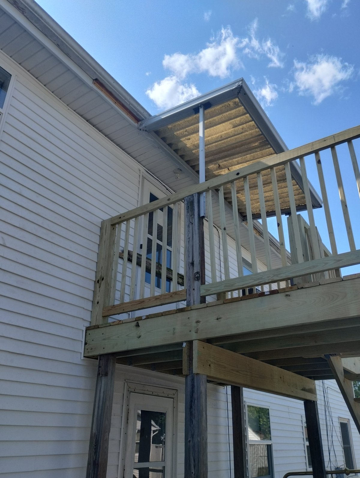 Two-story house with wooden deck and railing, white siding, and pitched roof against blue sky with clouds