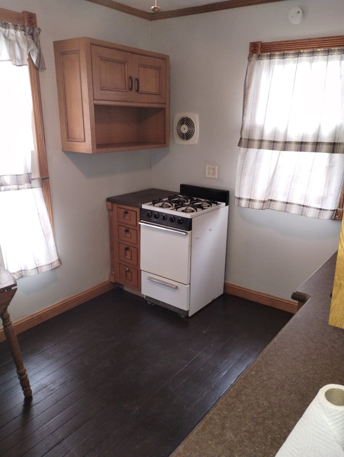 Compact kitchen with white gas stove, wooden cabinets and shelving, dark wood flooring, and windows with checkered curtains