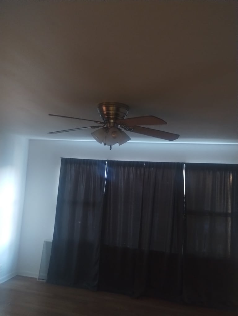 Bronze ceiling fan with blades mounted on white ceiling above dark curtains and hardwood floor in a room