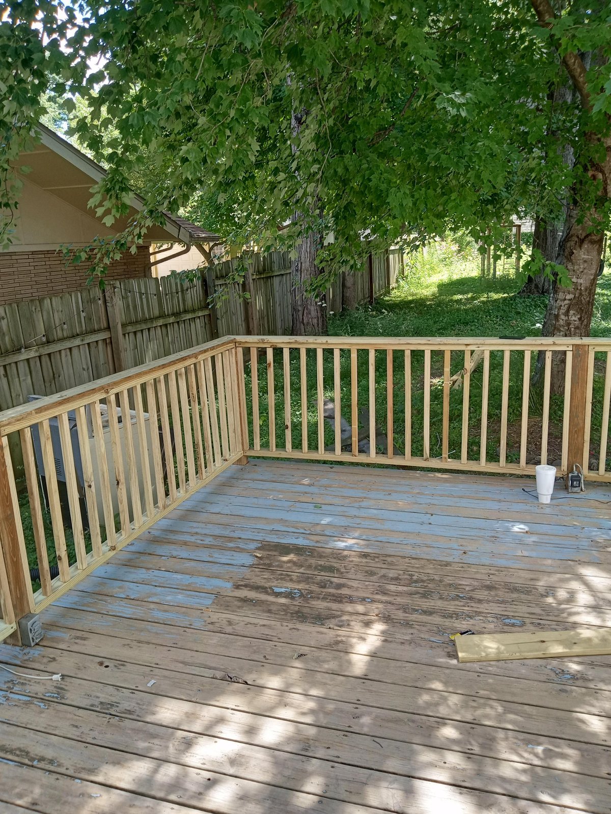 Wooden deck with light brown railings under large shade trees, partially weathered boards visible in dappled sunlight