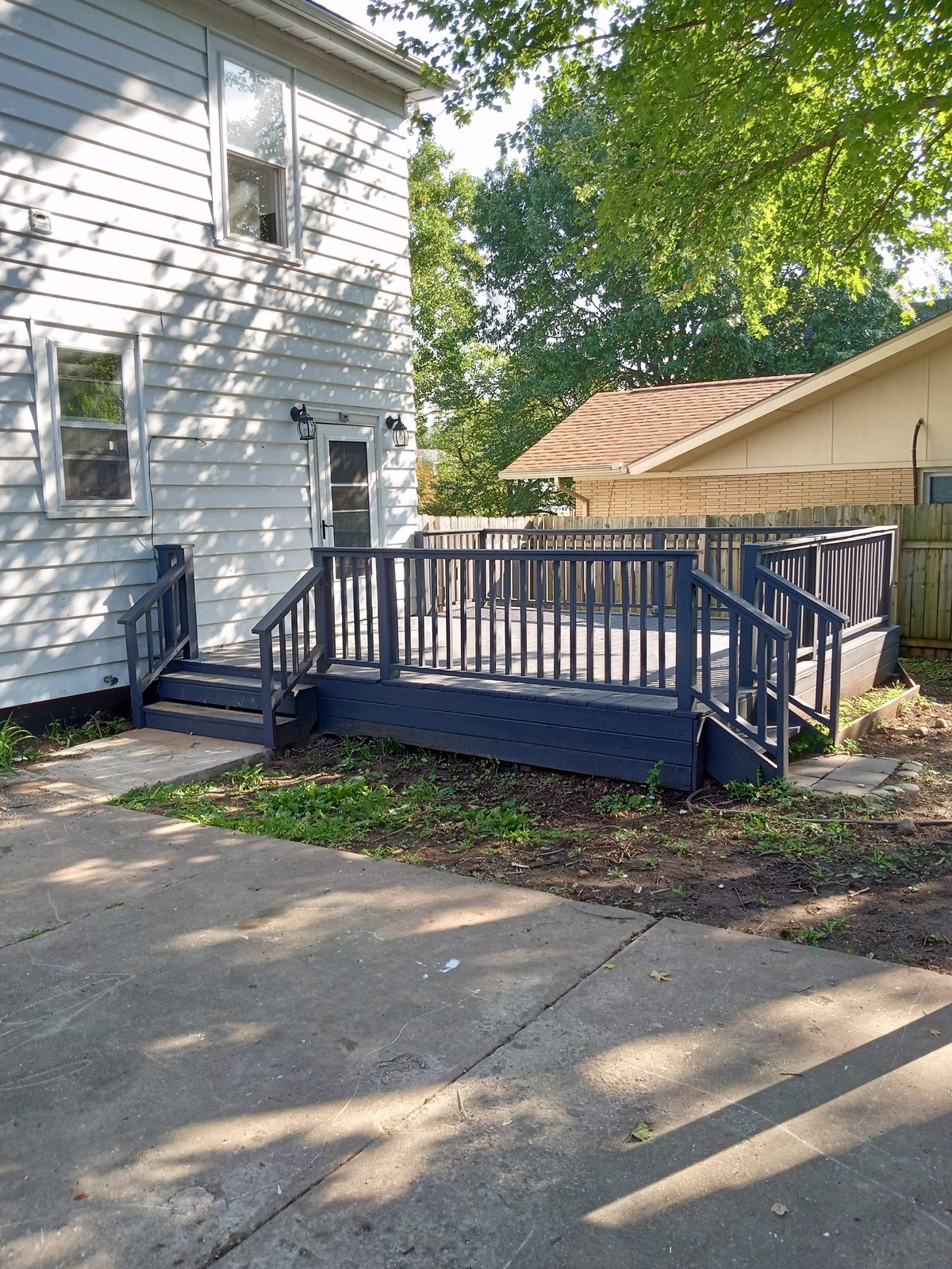 A dark blue composite deck with white railings attached to a light blue-gray house, surrounded by green trees and landscaping