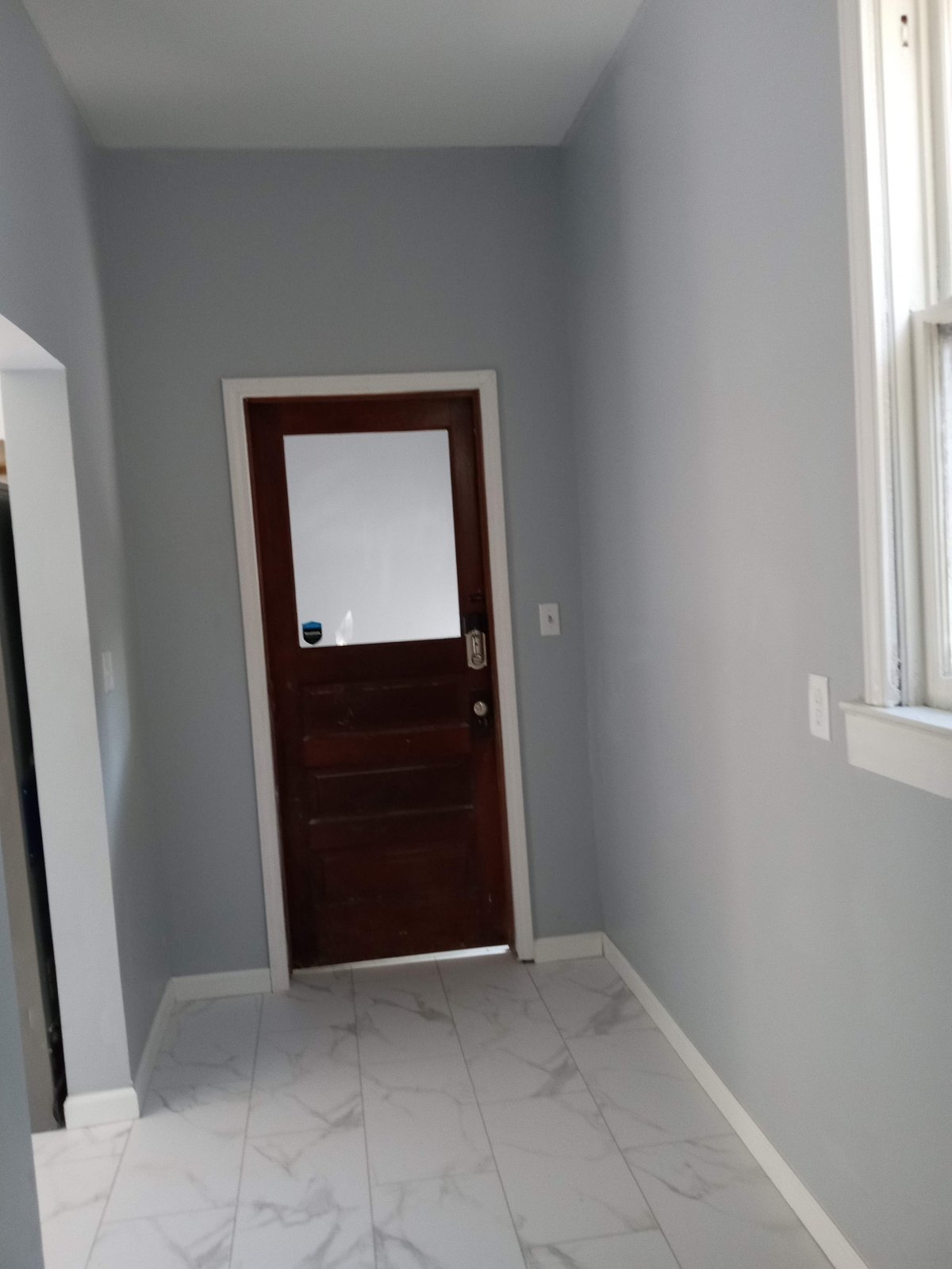 Small interior hallway with gray walls, white marble-look tile flooring, dark wooden door with window, and light from a window on the right