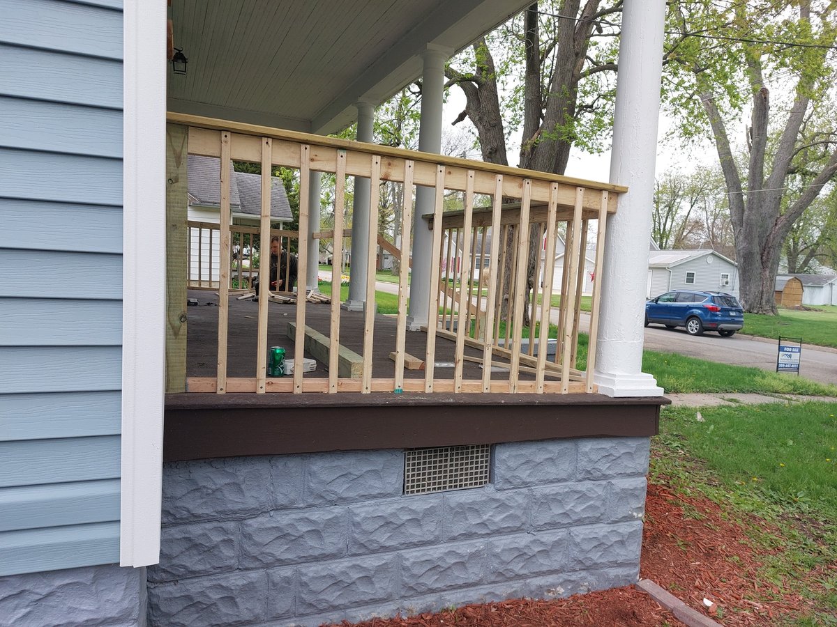 Covered porch with wooden railings and gray stone siding on residential house with trees and parked car visible in yard