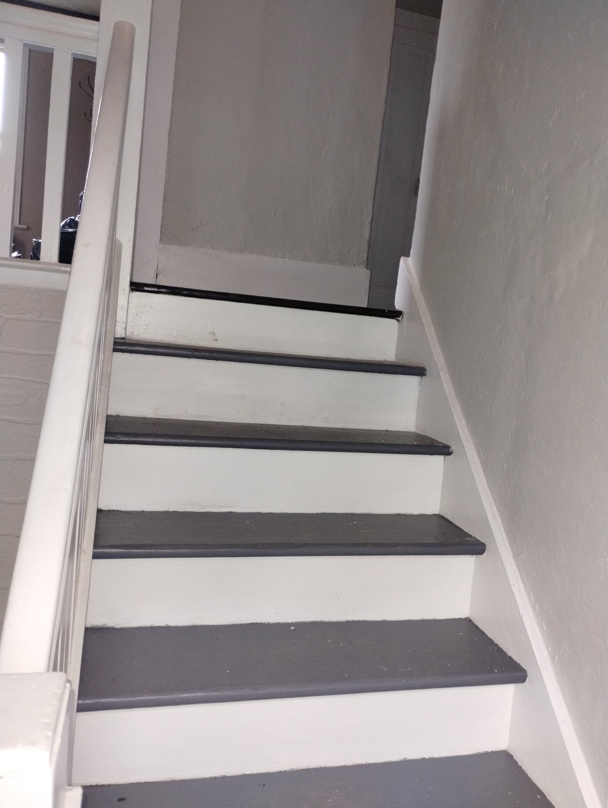 Interior staircase with white walls and alternating white and dark gray steps, viewed from bottom looking upward toward a landing with a door